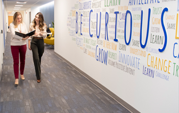 two women walking in front of a wall that says be curious